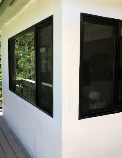 A corner of a white building with black-framed windows overlooking a porch with greenery in the background.