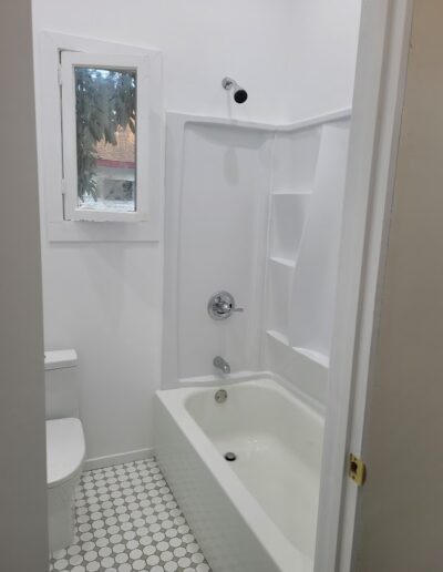 A small bathroom with a white toilet and bathtub, hexagonal floor tiles, and a window with a view of trees.