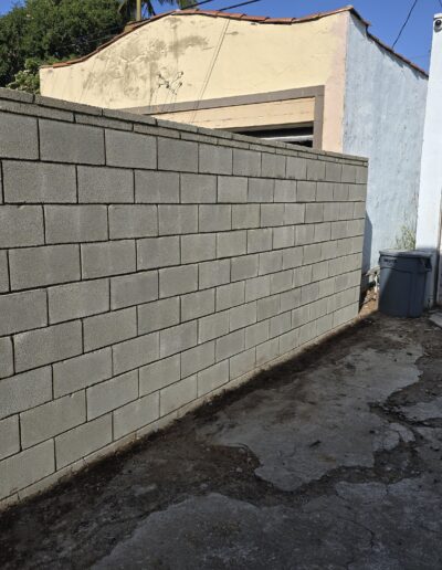 Concrete block wall next to a garage with a white door and a gray trash bin, set on cracked pavement.