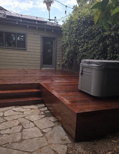 Wooden deck with steps leads to a beige house, featuring a large gray hot tub. Surrounded by greenery and string lights above.