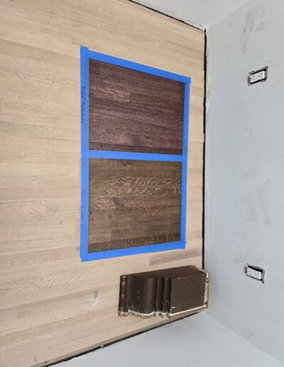 Two wood stain samples on a floor, enclosed in blue tape, with paint swatches leaning against a wall in a room under renovation.