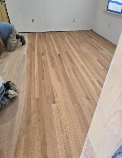 Two workers are installing light-colored wooden flooring in an empty room with white walls and a window.