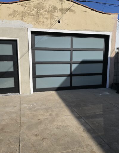 Modern garage with a frosted glass door and an adjacent matching entry door. The concrete driveway is partially in shadow.