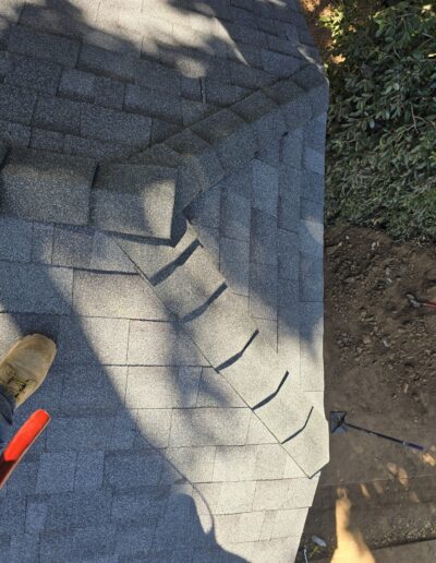 A rooftop with gray asphalt shingles and shadows cast over it. A small section of tools and boots are visible in the corner.