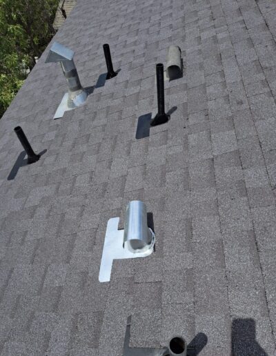 Gray shingled roof with multiple vents and stacks. Some vents have metal coverings. Sparse greenery is visible in the background.