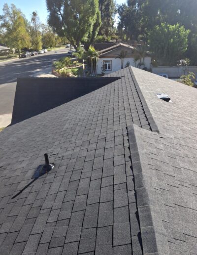 View of a residential rooftop with new asphalt shingles, chimney pipe, and a nearby neighborhood street.