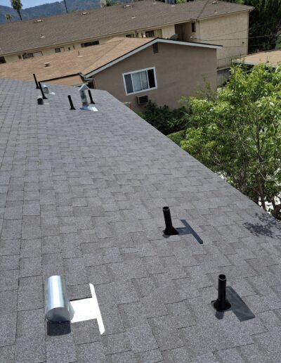 Several roof vents are installed on a shingled roof, with residential buildings and trees visible in the background.