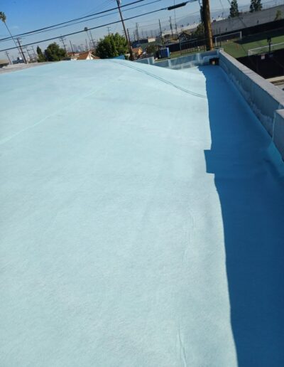 A flat rooftop covered in a light blue waterproof membrane under a clear sky, with surrounding buildings and a utility pole in the background.